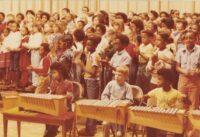 Three students sitting with musical instruments. A larger group of students stand behind them clapping.