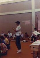 One teachers stands behind a group of adults, possibly conducting music.