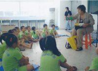 Teacher sits amongst a group of school children holding stuffed animals.