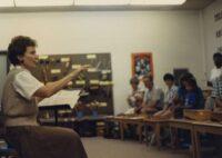 One teacher sits in front of a group of school children holding a musical score with her hand in the air conducting.